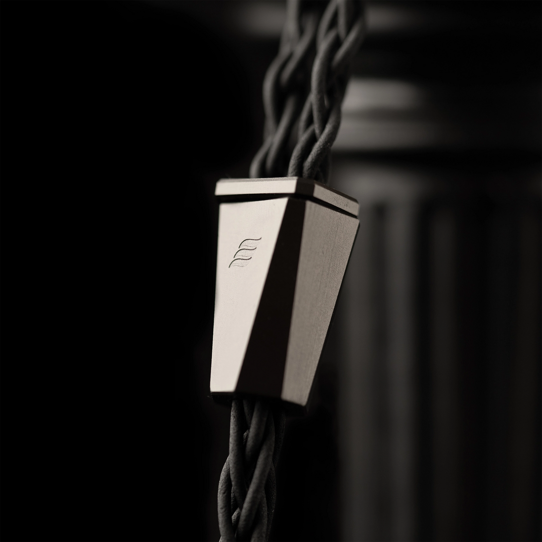 A close-up of Effect Audio Perseus iem metallic silver geometric cable connector with a brushed finish, attached to thick black braided cords against a dark, moody background.